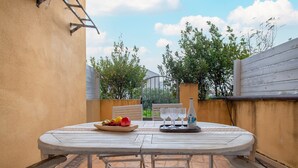 Un tavolo esterno rotondo con una ciotola di frutta e bicchieri da vino, sullo sfondo di un muro color beige e vista su alberi e un cancello.