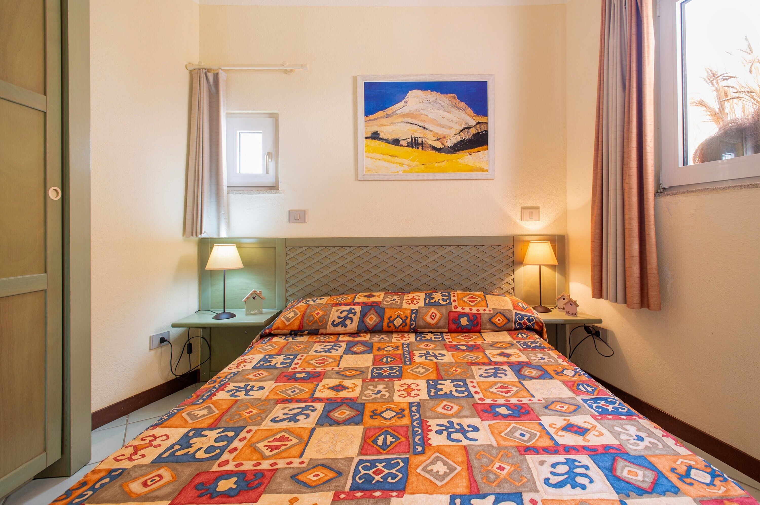 Une chambre d’hôtel avec un lit, deux lampes de chevet, un tableau au mur et une fenêtre avec des rideaux.
