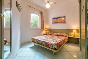 Une chambre à coucher comprenant un lit, deux tables de chevet, un ventilateur de plafond, une fenêtre et un tableau accroché au mur.
