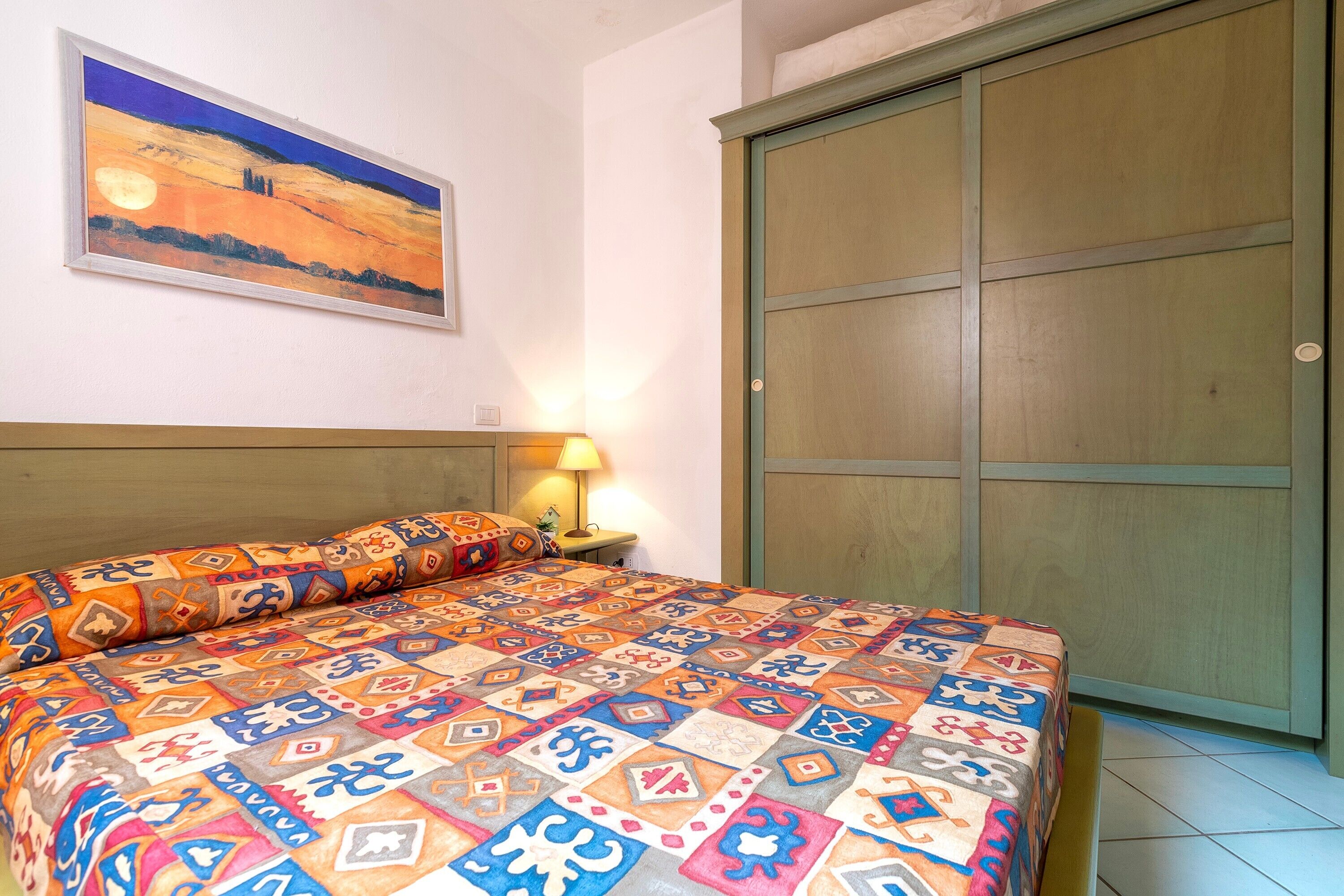 A bed with a patterned bedspread, a nightstand with a lamp, a painting on the wall, and a wardrobe.