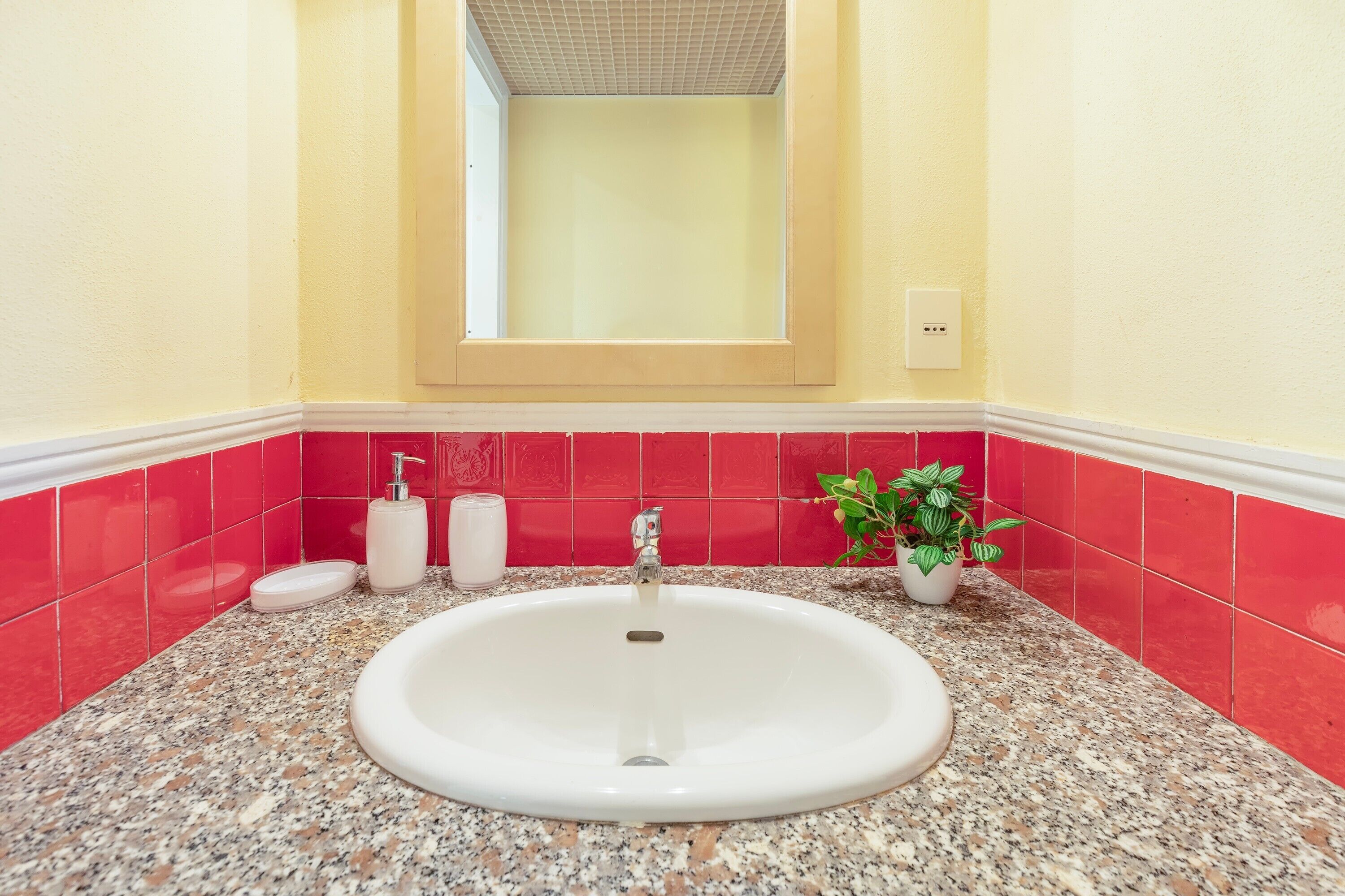 A bathroom with a white sink, red tiles, a potted plant, and a window above the sink.