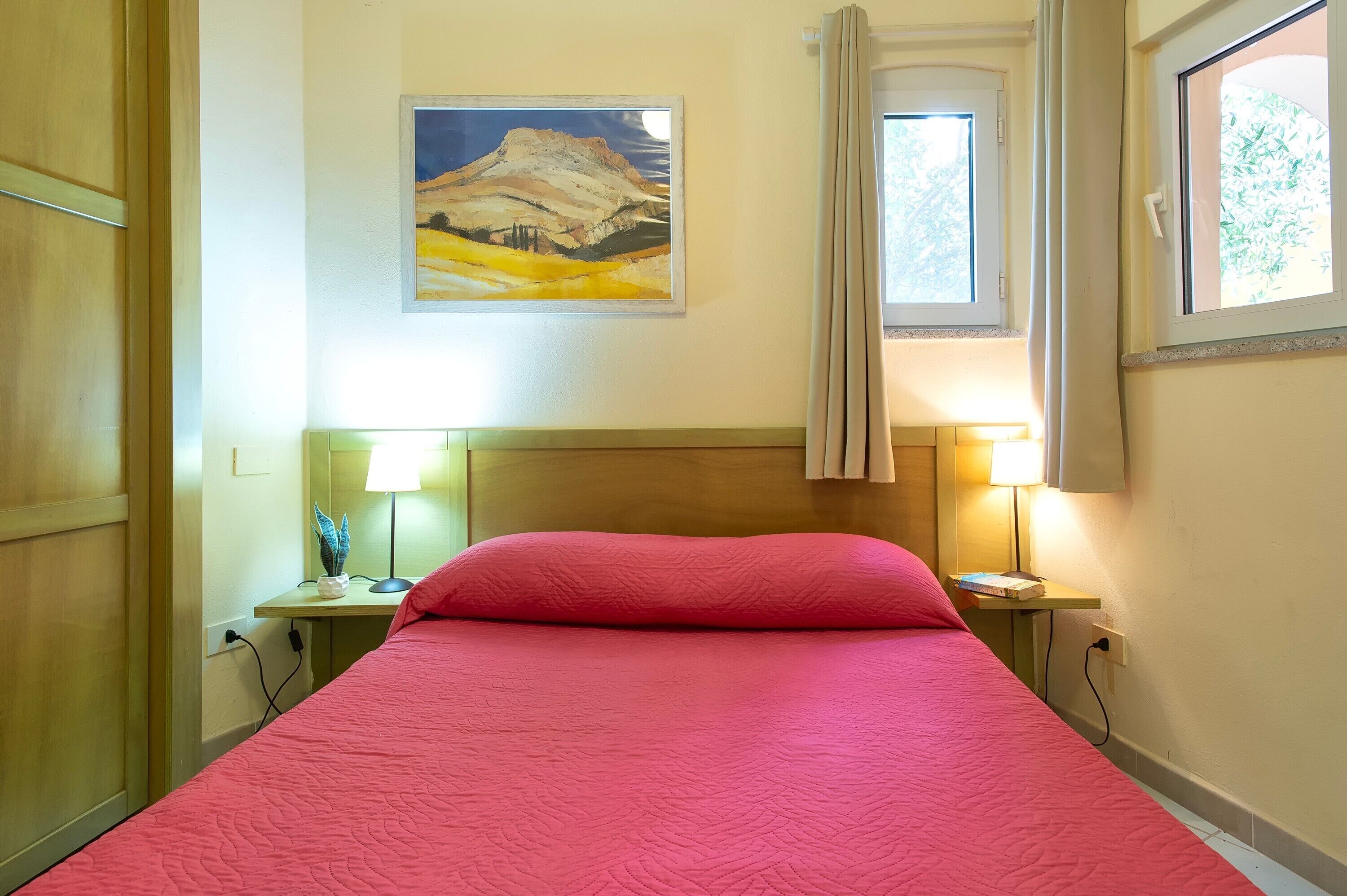A bed with a pink bedspread, a painting of a mountain landscape on the wall, and two bedside lamps.