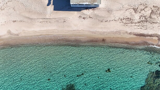 An aerial view of a beach with clear turquoise water, a sandy shore, and a structure in the background.