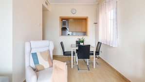 A living room with a sofa, a dining table with chairs, a kitchen area with cabinets, and a clock on the wall.