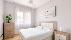 A bedroom with a bed, a nightstand, a dresser, a ceiling fan, a window with curtains, and a wall-mounted artwork.