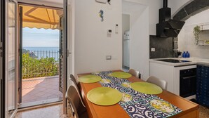 An open door leading to a balcony with a view, a dining table with placemats, a kitchen with a stove, and a hanging light fixture. - Apartment 'Profumo Di Salsedine' with Sea View, Wi-Fi and Air Conditioning (Bonassola)
