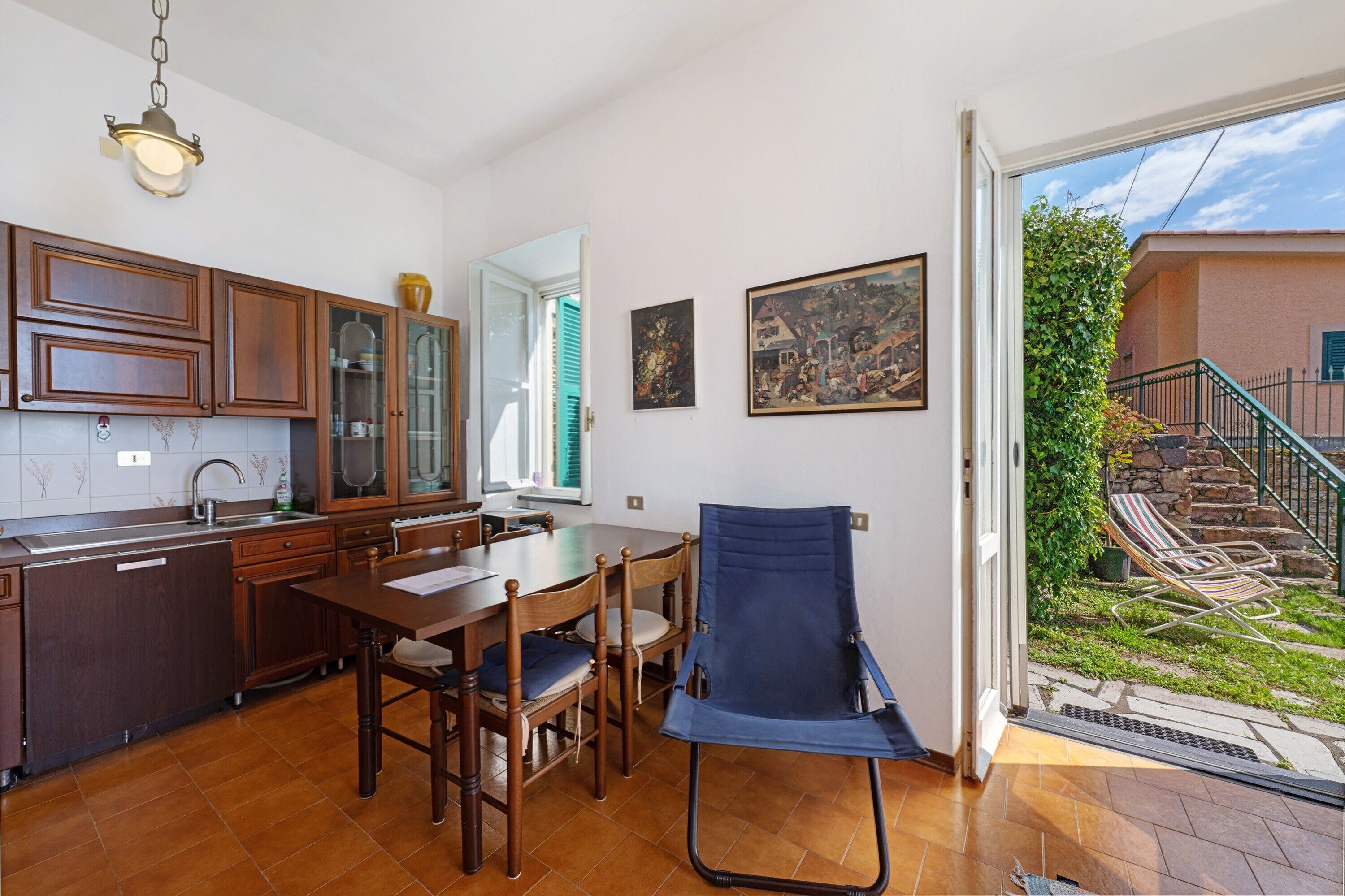 A kitchen with wooden cabinets, a dining table with chairs, a blue folding chair, and a view of a garden through an open door.