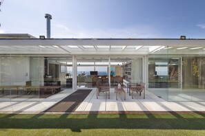 A modern house with a glass facade, outdoor seating, and a view of the sea.