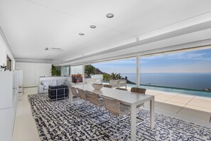 A modern kitchen with white cabinets, a dining area with a large table and chairs, and a view of the ocean.