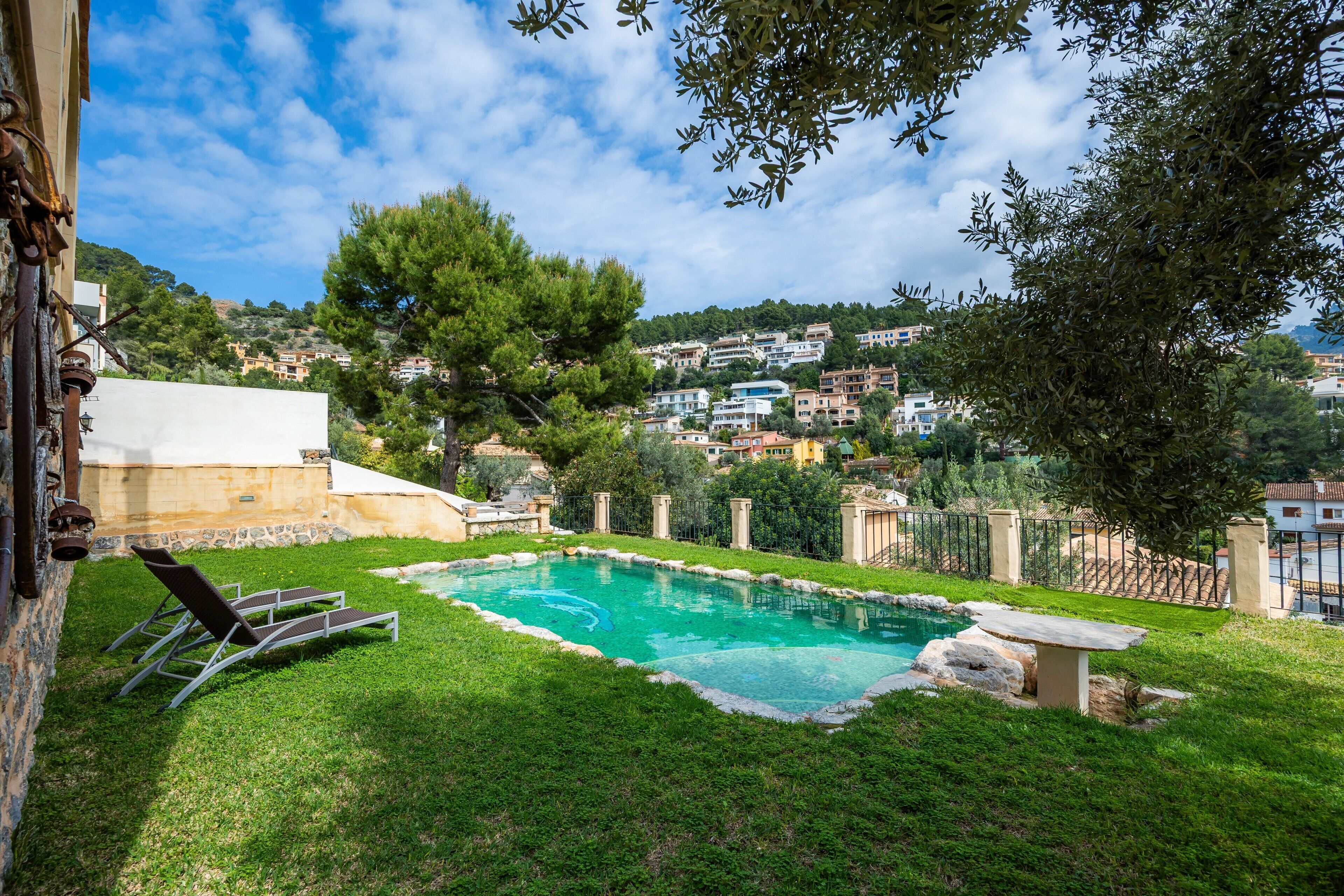 A swimming pool with lounge chairs, surrounded by a grassy area and a stone wall, with a view of a hillside town in the background.