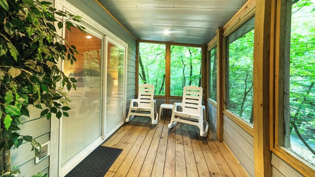 A covered porch with wooden flooring, two white plastic chairs, and large windows offering a view of a green forest. - Pam's Treehouse Cabin #1-A favorite treehouse right on a creek! (Hot Springs)