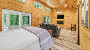 A cabin with a bed, a recliner, a TV, and large windows with a forest view. - Pam's Treehouse Cabin #1-A favorite treehouse right on a creek! (Hot Springs)