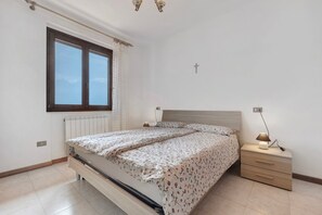Une chambre à coucher avec un lit, une table de chevet, une lampe, une fenêtre avec des rideaux et une croix au mur.