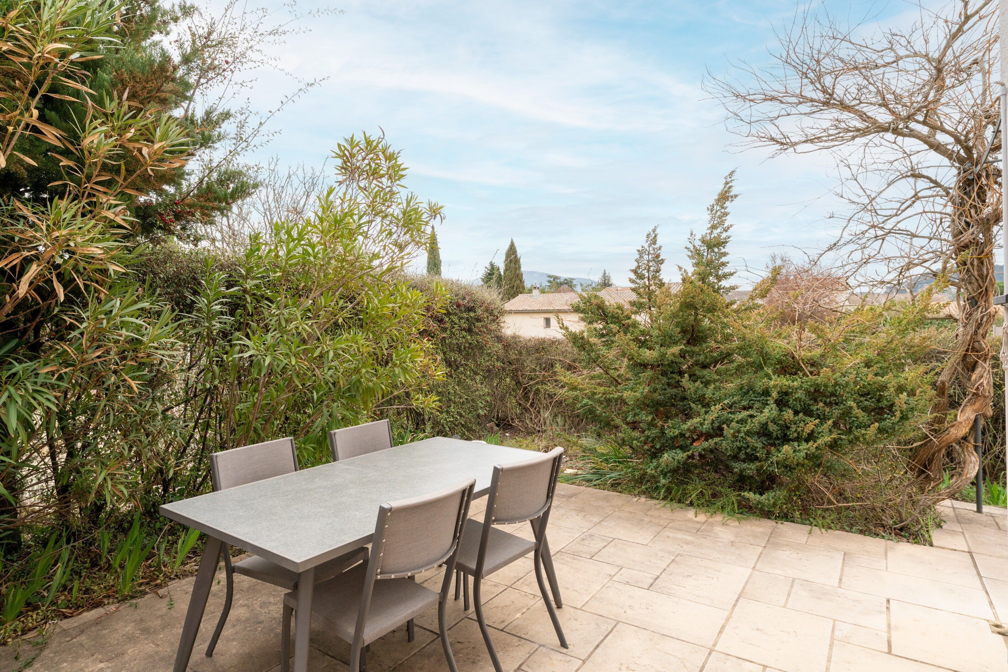 An outdoor patio with a table and chairs, surrounded by greenery and a view of a building in the distance.