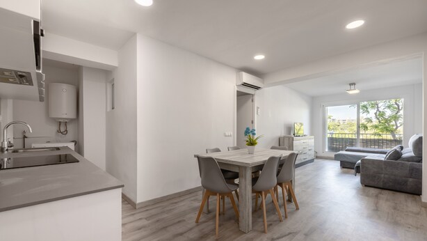 Una cocina moderna con horno integrado, un comedor con mesa y sillas de madera, un sofá gris y un amplio ventanal con vistas a zonas verdes.