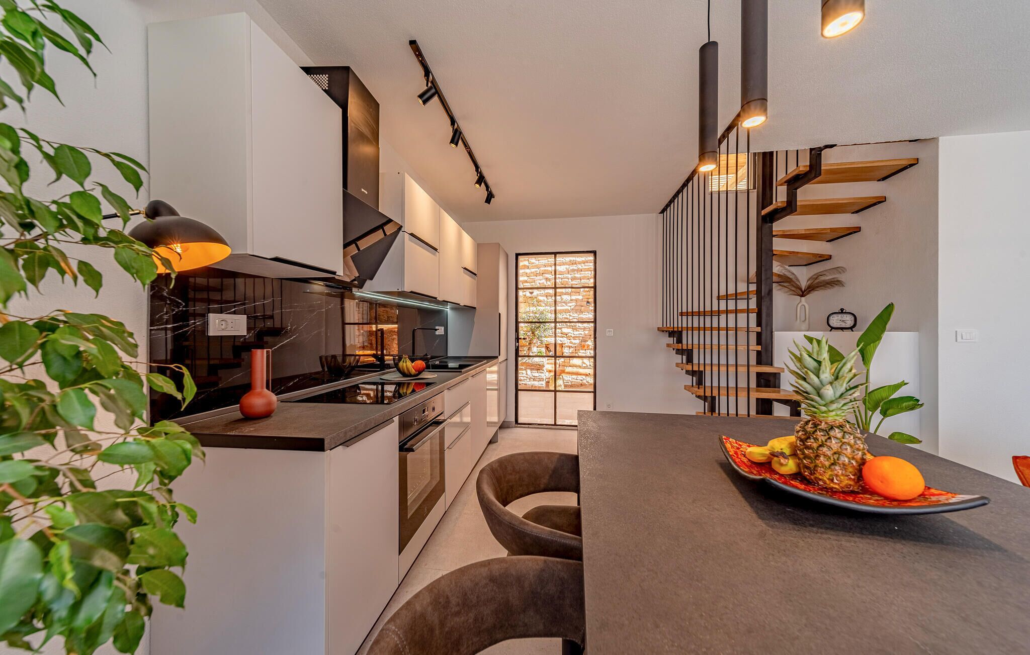 A modern kitchen with a central island, a dining area with a fruit bowl, and a staircase with a wooden finish.