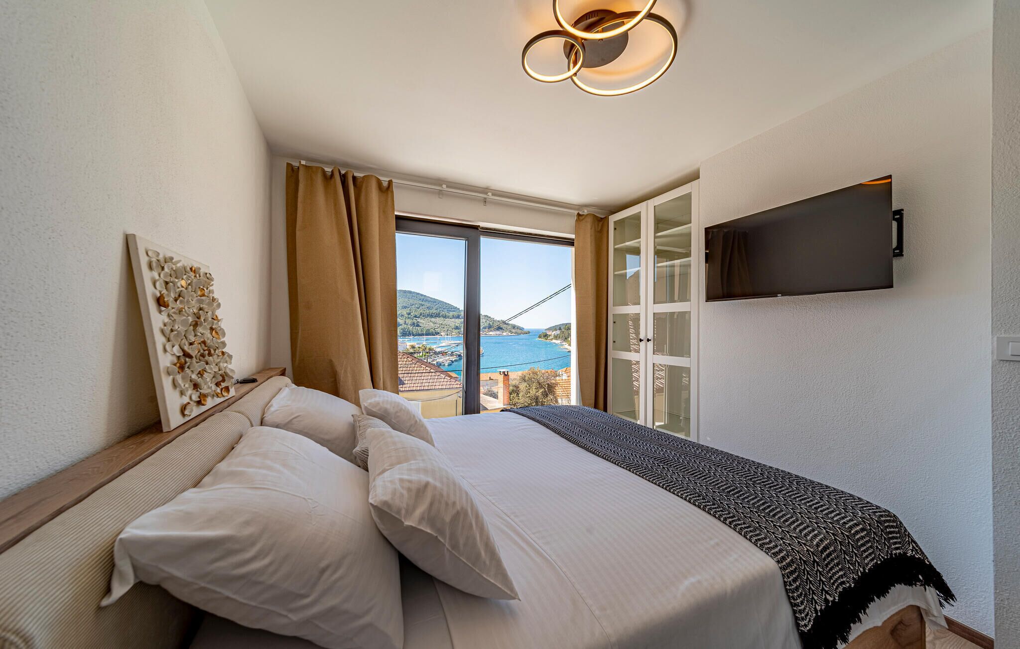 Habitación de hotel con cama, vista a la costa, televisor montado y lámpara en el techo.