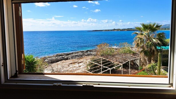 A window view of a coastal landscape with a clear sky, a rocky shoreline, and a palm tree. - VILLA THE HELM - ORTIGIA HOLIDAYS (Fontane Bianche)