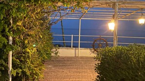 A brick pathway leading to a covered area with a large wheel structure, surrounded by greenery and illuminated by wall-mounted lights. - VILLA THE HELM - ORTIGIA HOLIDAYS (Fontane Bianche)