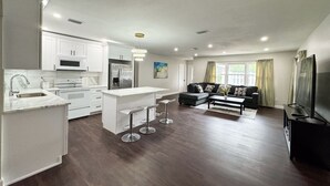 A modern kitchen with white cabinetry, a central island with stools, and a dining area with a black sofa and coffee table. - Home Close to Saint Pete Pier new Spacious (St. Petersburg)