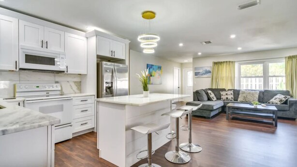 A modern kitchen with white cabinetry, a central island with stools, and a dining area with a sofa and coffee table. - Home Close to Saint Pete Pier new Spacious (St. Petersburg)