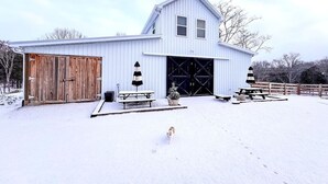 Ein weißes Bauernhaus mit einer großen Garagentür, einem Holzschuppen, Picknicktischen und einem mit Schnee bedeckten Boden.
