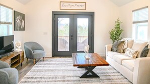 Ein Wohnzimmer mit einer dunklen Tür, einem Fernseher, einem grauen Sessel, einem Holztisch und einem beigen Sofa.