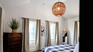 Ein Schlafzimmer mit karierter Bettdecke, einem hölzernen Schminktisch, einer hängenden Pflanze und einer gewebten Leuchte.