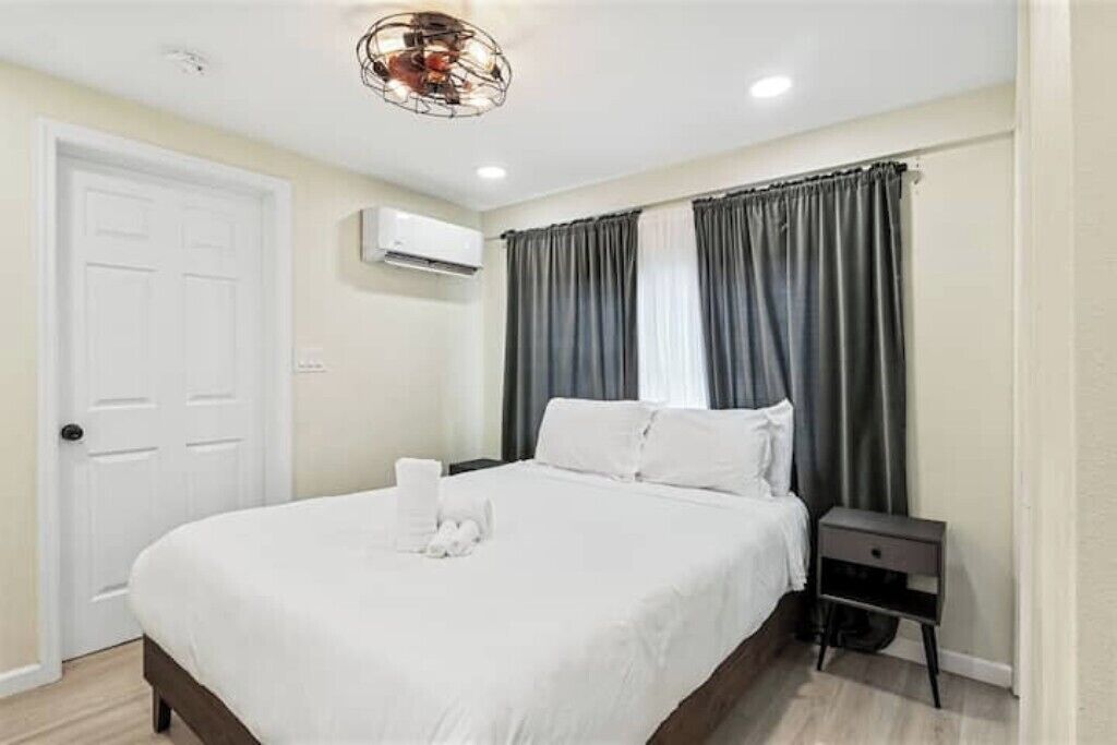 Una cama bien tendida con sábanas blancas, una mesita de noche, un aire acondicionado de pared y una puerta.