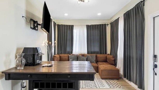A modern living room with a dark wooden coffee table, a flat-screen TV mounted on the wall, a brown leather sofa, and dark curtains.