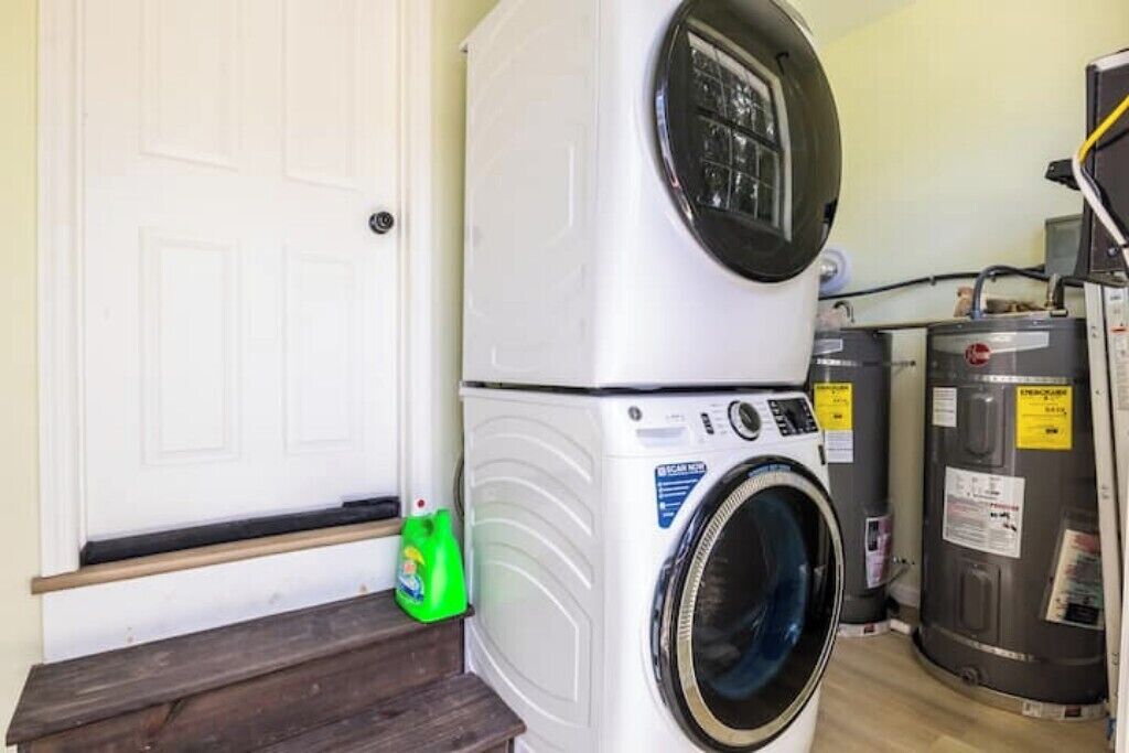 A stacked washer and dryer unit in a laundry room with a wooden bench and a green detergent bottle.