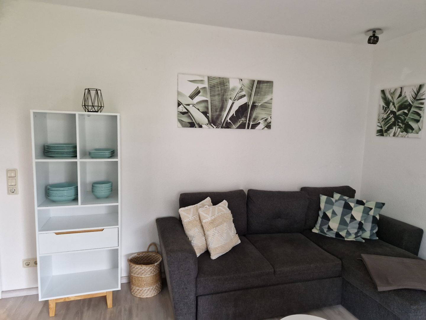 A living room with a grey sofa, a white shelving unit with teal plates, and a framed leaf artwork on the wall.