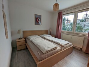 A bedroom with a wooden bed, two bedside tables, a hanging lamp, a window with curtains, and a radiator.