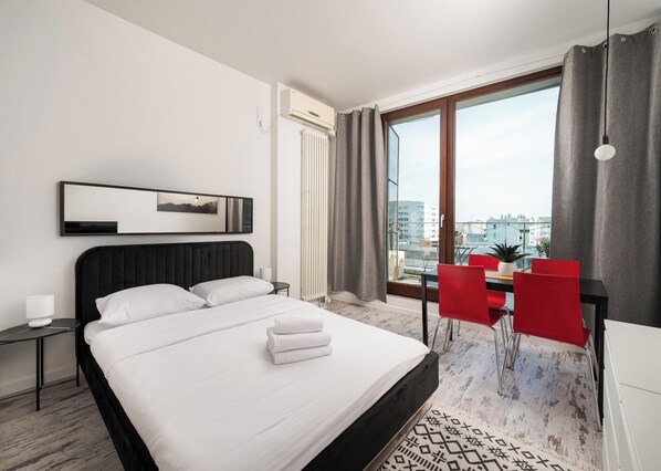 A modern hotel room with a large bed, a small table with a lamp, a chair, a view of the city through large windows, and a ceiling fan. - Ochota City View Stylish Studio (Warszawa)