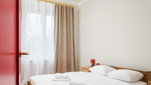 A neatly made bed with white linens and two folded towels, a wooden headboard, a small bedside table with a red lamp, and a window with beige curtains. - Metro Młynów Colourful Apartment (Warszawa)