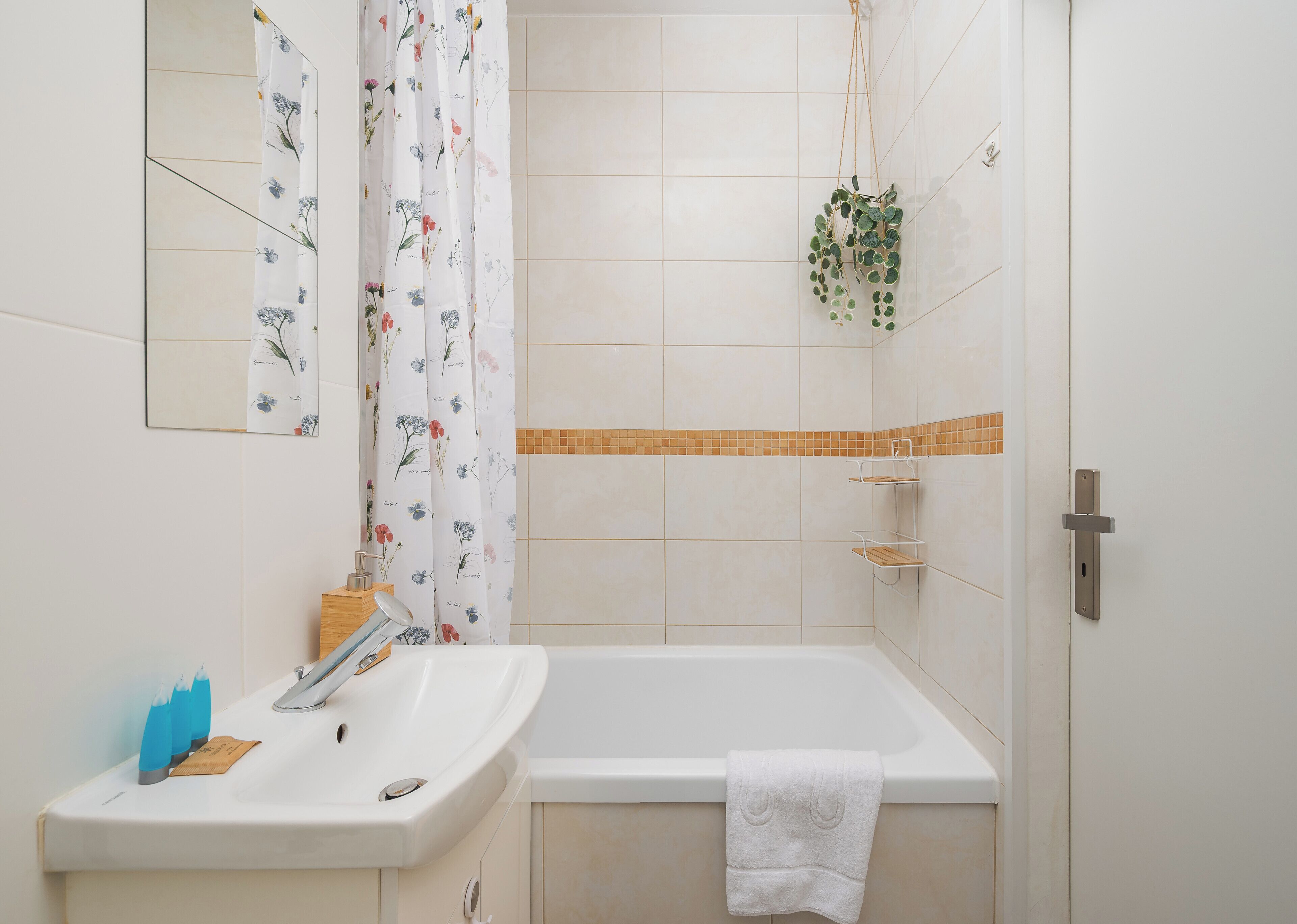 A bathroom with a white sink, a mirror, a shower curtain with floral patterns, a hanging plant, and a towel on the bathtub.