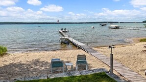 Ein Strand mit einer hölzernen Pier, die sich in ruhiges Wasser erstreckt, einer Flagge und einer Reihe von Stühlen.