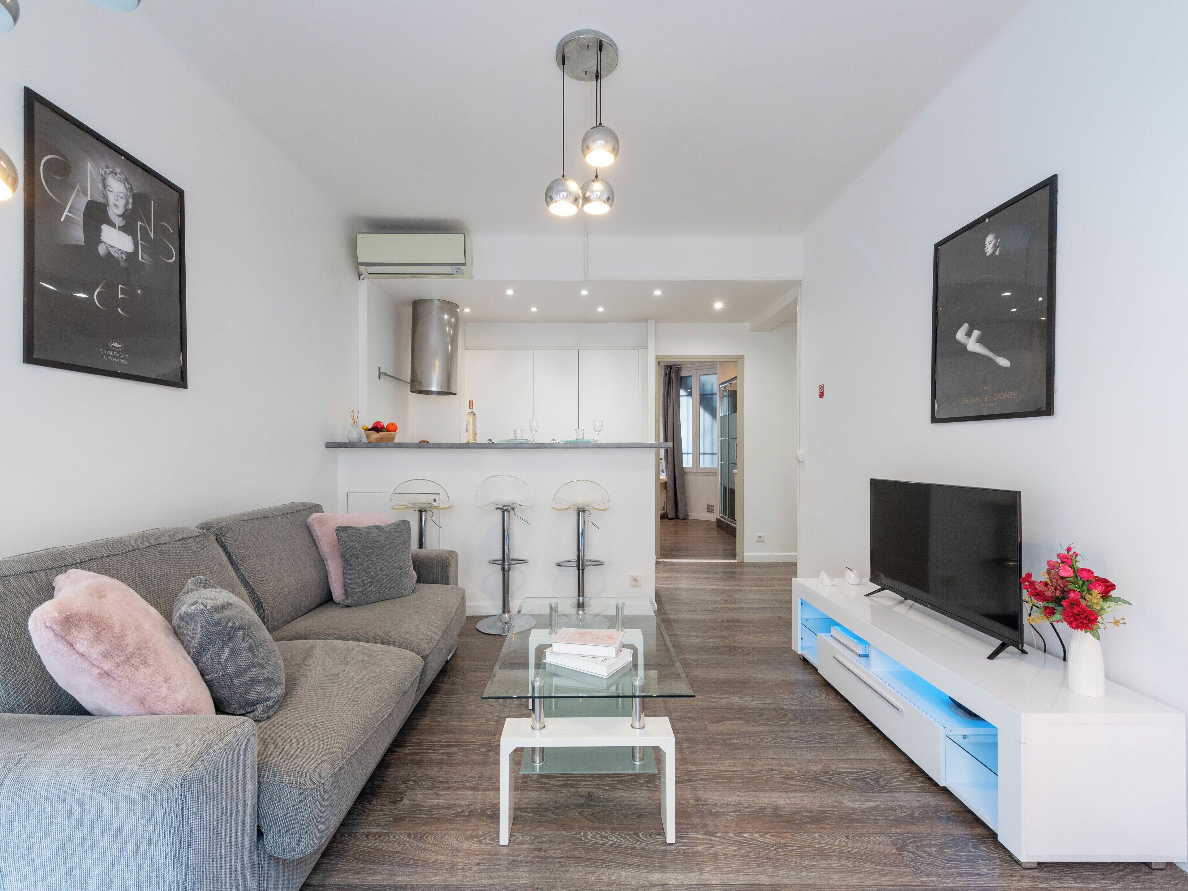 A modern living room with a grey sofa, a glass coffee table, a flat-screen TV, and a kitchen in the background.