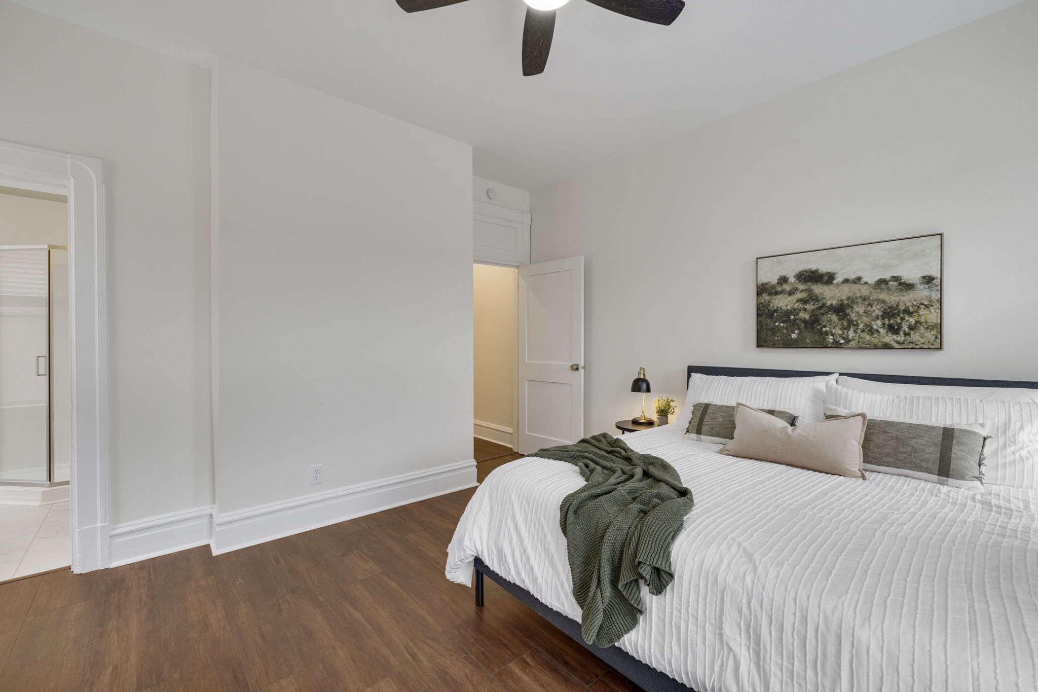 A bedroom with a bed, a ceiling fan, a nightstand with a lamp, and a painting on the wall.