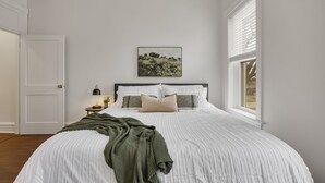 A bedroom with a large bed, a bedside table with a lamp, a framed picture on the wall, and a window with blinds. - 3BR Historic St. Charles, MO-Walking distance to Main Street (Saint Charles)