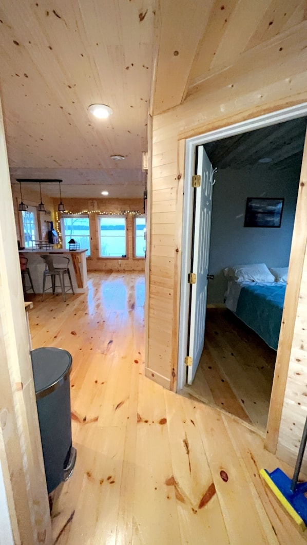A wooden interior with a kitchen area, a dining space, and a bedroom with a bed and a painting.