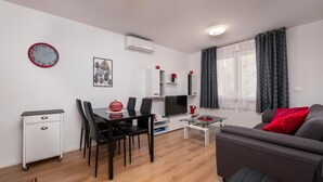 A modern living room with a grey sofa, a dining area with a glass table and black chairs, a flat-screen TV, and a clock on the wall. - Apartman Porec Center Sea (Porec)