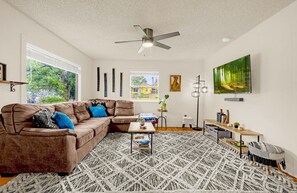 A living room with a brown leather sofa, a patterned rug, a ceiling fan, a TV, and a window with a view of a house.