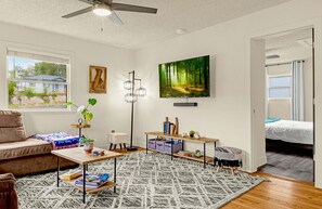 En stue med sofa, sofabord, gulvlampe, fjernsyn og udsigt til et hus.