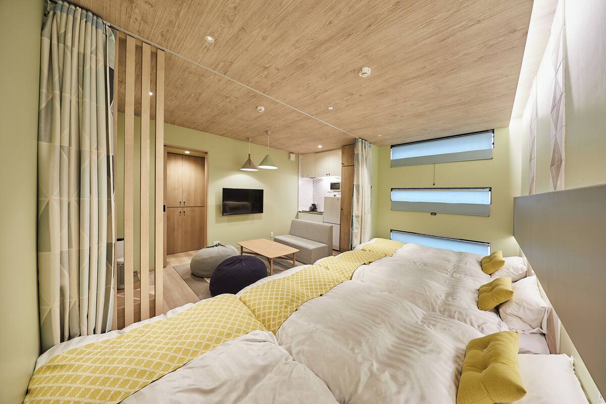 A modern bedroom with a large bed, a desk, and a kitchenette in the background.