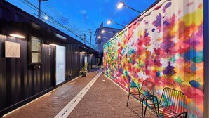 A narrow alleyway with a colorful mural on one side and a black building on the other. There are two metal chairs outside the building. - TORACO konohana (Osaka)