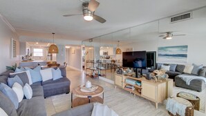 Ein geräumiges Wohnzimmer mit einer großen, grauen Couch, einem Holztisch und einem Fernseher. Im Hintergrund befindet sich eine Küchenzeile mit einem Esstisch und Stühlen.