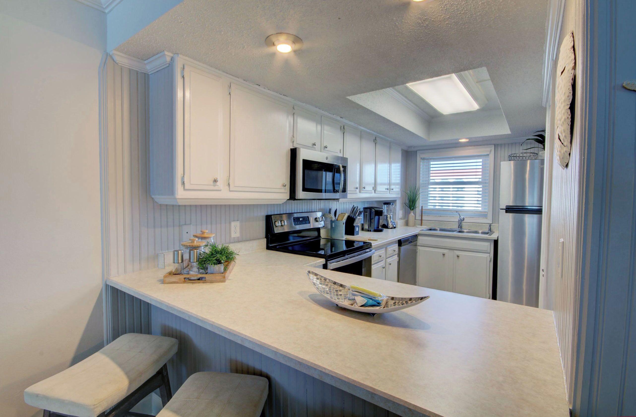 A modern kitchen with white cabinetry, a microwave, stove, and a refrigerator. There are two bar stools and a decorative bowl on the counter.