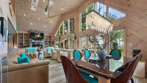 Amplia sala de estar con techo de madera, ventanas grandes, chimenea y una mesa de comedor con sillas.