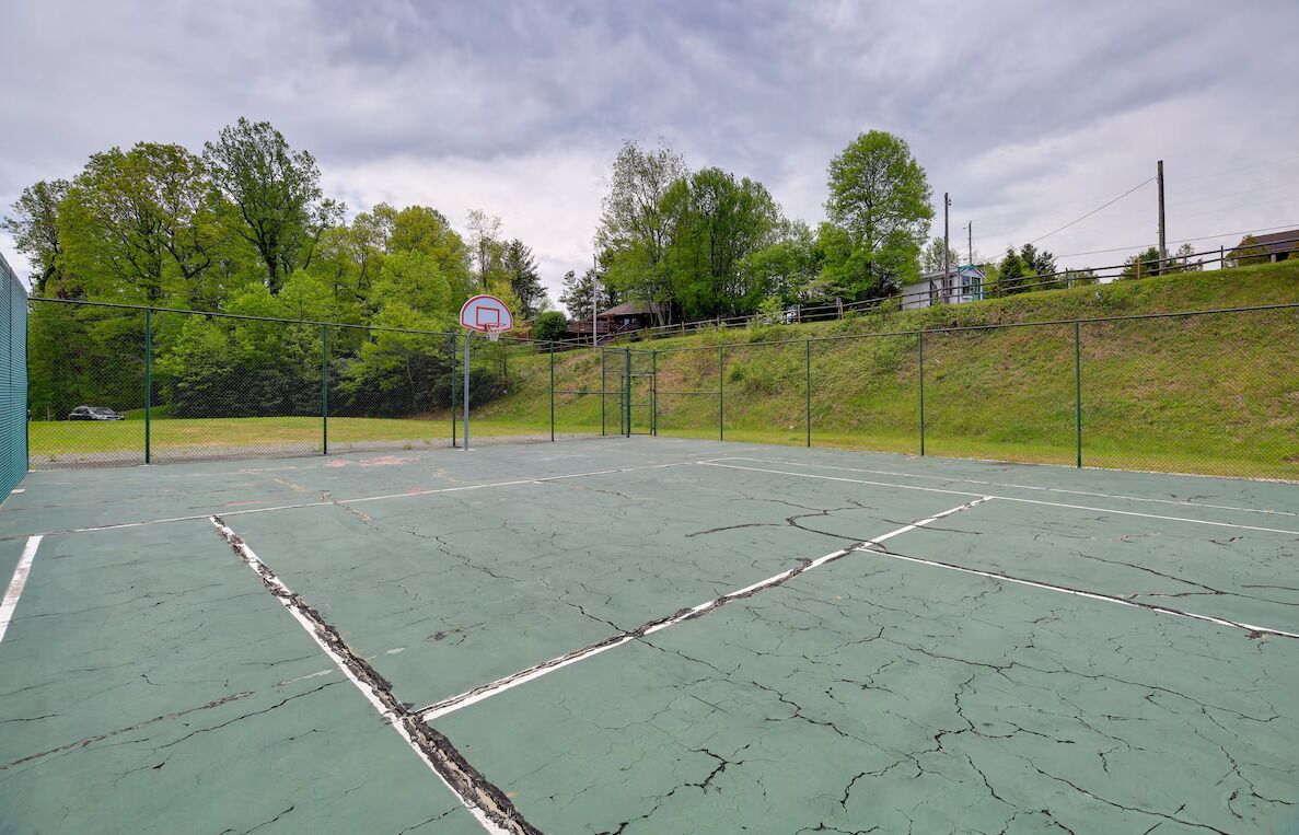 Una cancha de baloncesto al aire libre con superficie verde, rodeada por una cerca de malla metálica, árboles y un edificio al fondo.